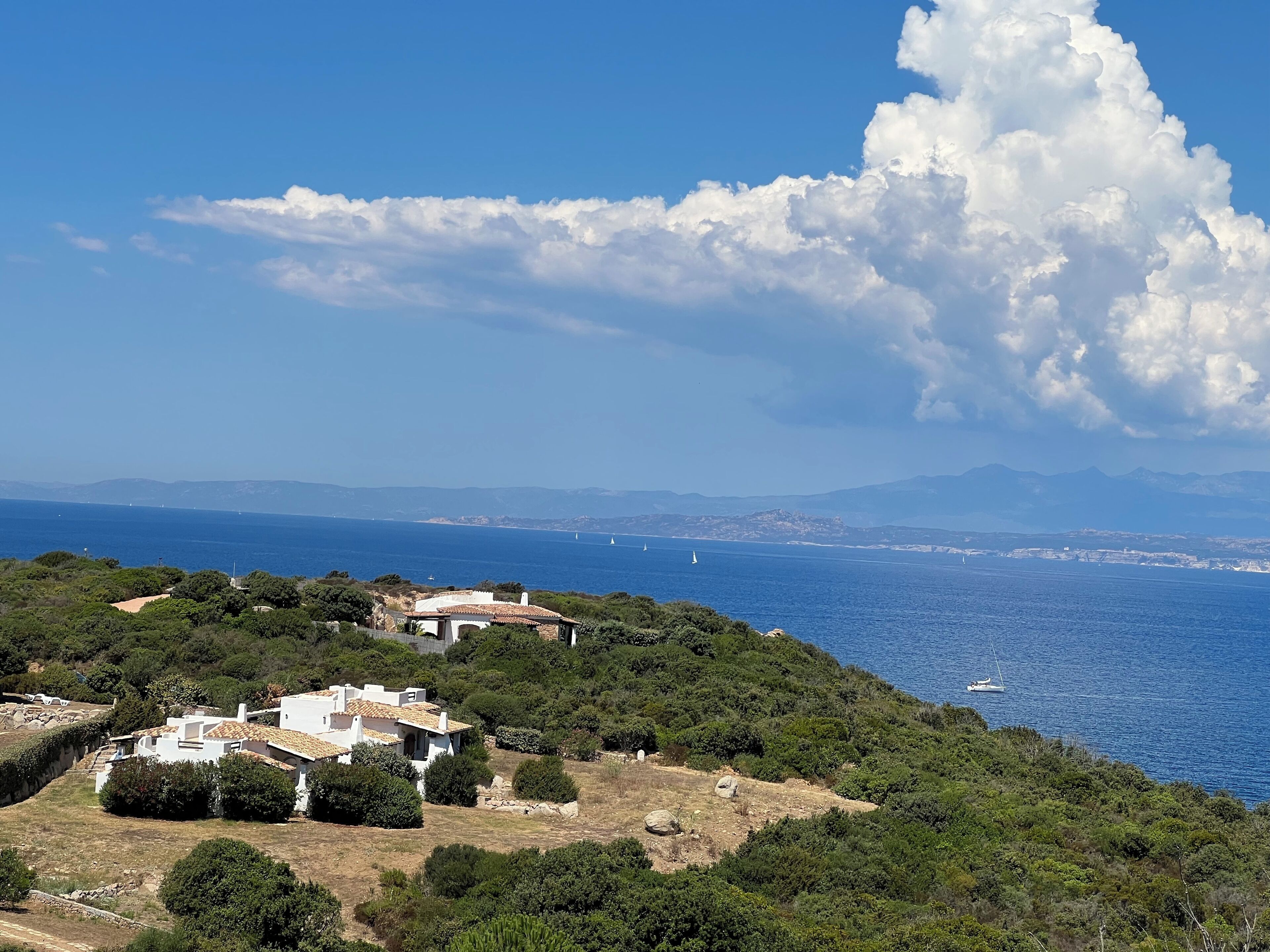 A coastal landscape with a white building surrounded by greenery, overlooking the sea with sailboats and distant mountains.