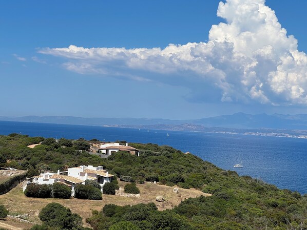 A coastal landscape with a white building surrounded by greenery, overlooking the sea with sailboats and distant mountains.