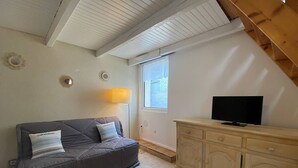 Un salon chaleureux avec un canapé gris, un téléviseur posé sur un meuble en bois et une fenêtre donnant sur un mur de briques.