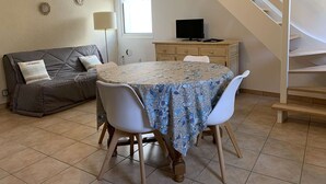 Un salon chaleureux avec une table ronde, deux chaises blanches, un canapé gris, un téléviseur posé sur un meuble en bois et un escalier.
