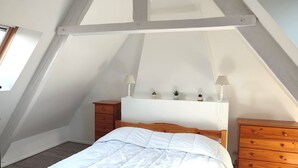 Une chambre avec un plafond en pente, un lit en bois, une table de chevet, une commode et une étagère ornée d’objets décoratifs.