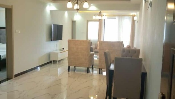 A spacious living area with a marble floor, a dining table with chairs, a sofa, and a TV mounted on the wall. - Stay.Plus Sandalwood Apartment Kilimani (Nairobi)
