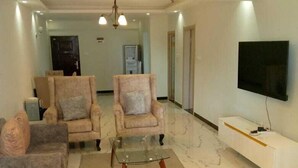 A modern living room with a grey sofa, two armchairs, a glass coffee table, and a flat-screen TV mounted on the wall. - Stay.Plus Sandalwood Apartment Kilimani (Nairobi)