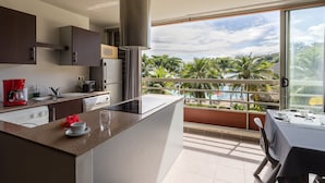 Une cuisine moderne dotée d’un grand îlot central, d’un coin repas avec une table et d’un balcon offrant une vue sur des palmiers et une plage.