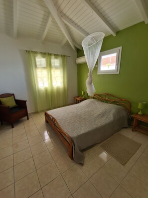 A bedroom with a bed, a chair, a window with curtains, a wall-mounted air conditioner, and a small table with a lamp.