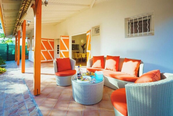 A covered outdoor seating area with wicker furniture and orange cushions, a dining area in the background, and a view of the outdoors.