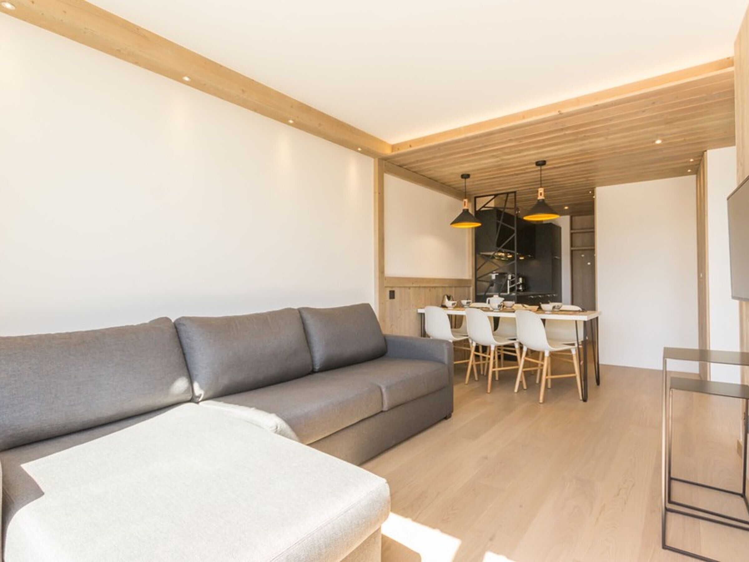 A modern living room with a grey sofa, a small table, and a dining area with chairs and a kitchen visible in the background.