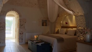 Een slaapkamer met een stenen plafond, een bed met witte lakens, een nachtkastje met kaarsen en een uitzicht door een boogvormige deuropening.