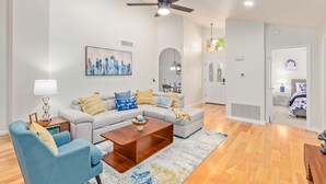 Ein modernes Wohnzimmer mit grauem Sofa, blauem Sessel und Couchtisch aus Holz. Man sieht einen Deckenventilator und ein Stadtbildgemälde.