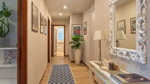 A hallway with a patterned rug, framed artwork, a potted plant, a console table with a lamp, and a mirror with an ornate frame. - Ampio appartamento vicino al mare con una grande terrazza solarium. (Alassio)