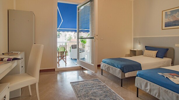 A hotel room with two beds, a desk, and a chair. There is a balcony with a view of the outside. - Ampio appartamento vicino al mare con una grande terrazza solarium. (Alassio)