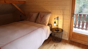 A cozy bedroom with a wooden ceiling, a bed with a white bedspread, two pillows with deer designs, a round side table with a light, and a balcony with a wooden railing. - Spacious chalet near shops, pets allowed! (Samoëns)