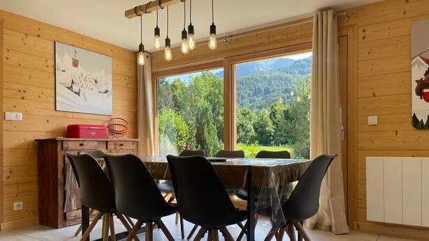 A dining area with a wooden table and chairs, a window with a view of greenery, and a ceiling light fixture. - Spacious chalet near shops, pets allowed! (Samoëns)