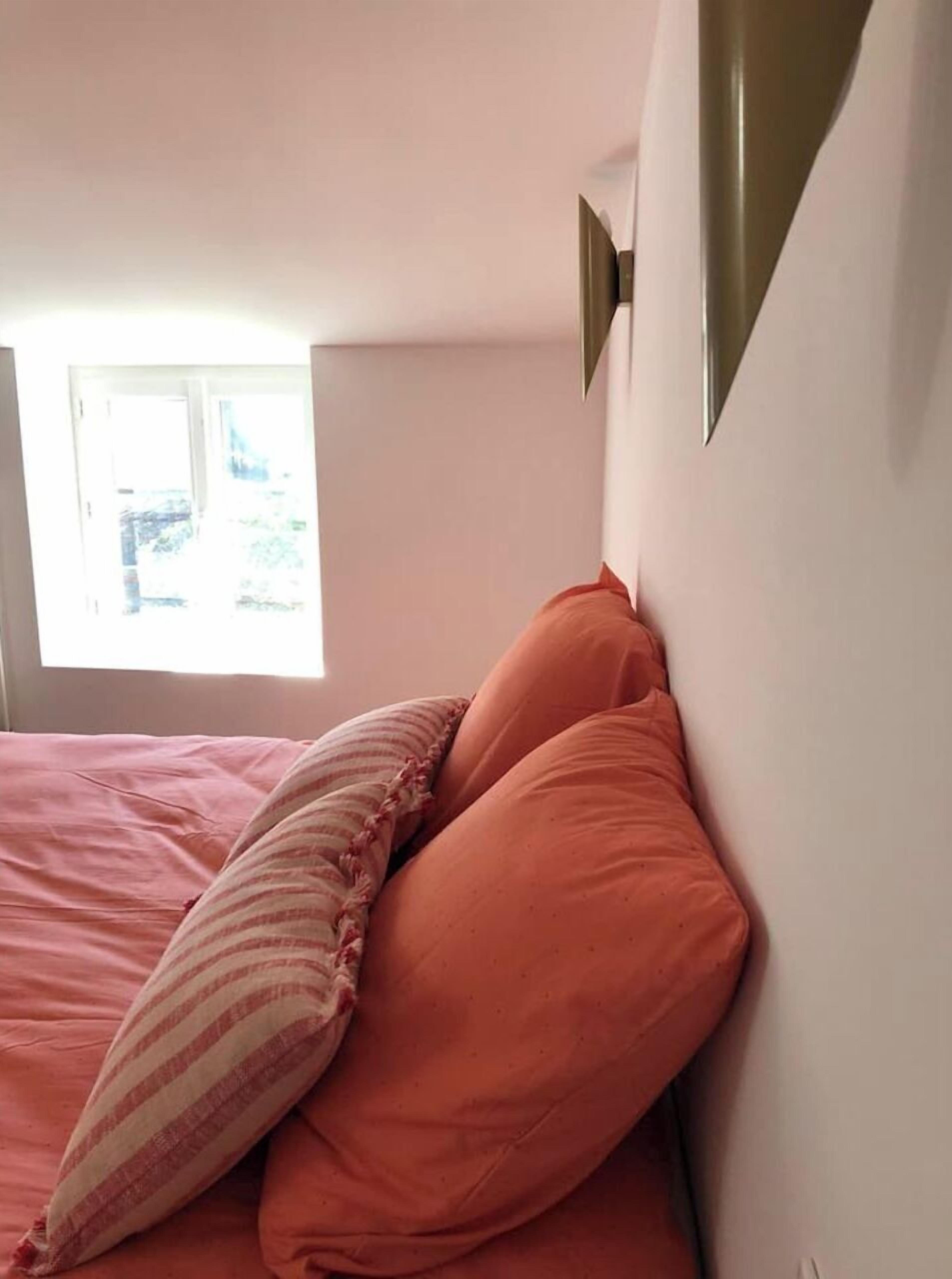 Une chambre à coucher avec un lit, des oreillers, une fenêtre et un luminaire fixé au mur.