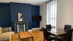 A modern living room with a fireplace, a glass dining table, and a large window with blinds. - Charming 1-bed Apartment in Lytham Saint Annes (Lytham St. Anne's)