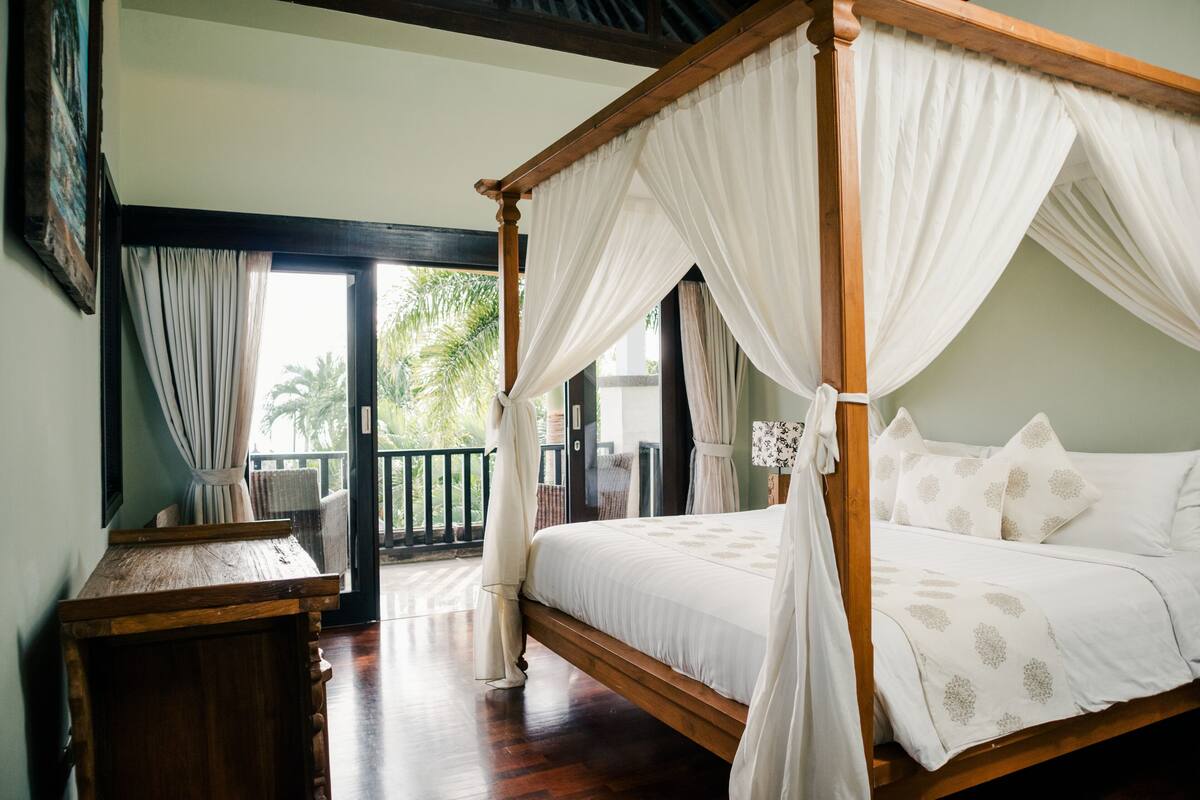 A four-poster bed with white bedding and a wooden frame, a wooden nightstand, and a view of the outdoors through a glass door.