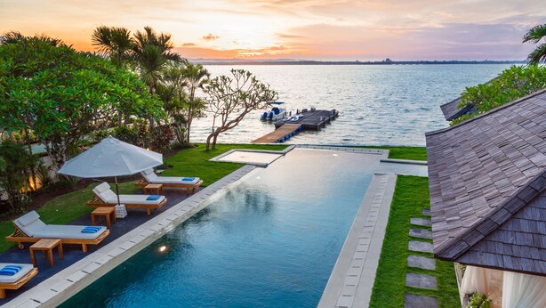 A swimming pool with lounge chairs and a dock extending into the water, surrounded by greenery and a view of the sunset. - Sunset Villa by Premier Hospitality Asia (Nusa Dua)