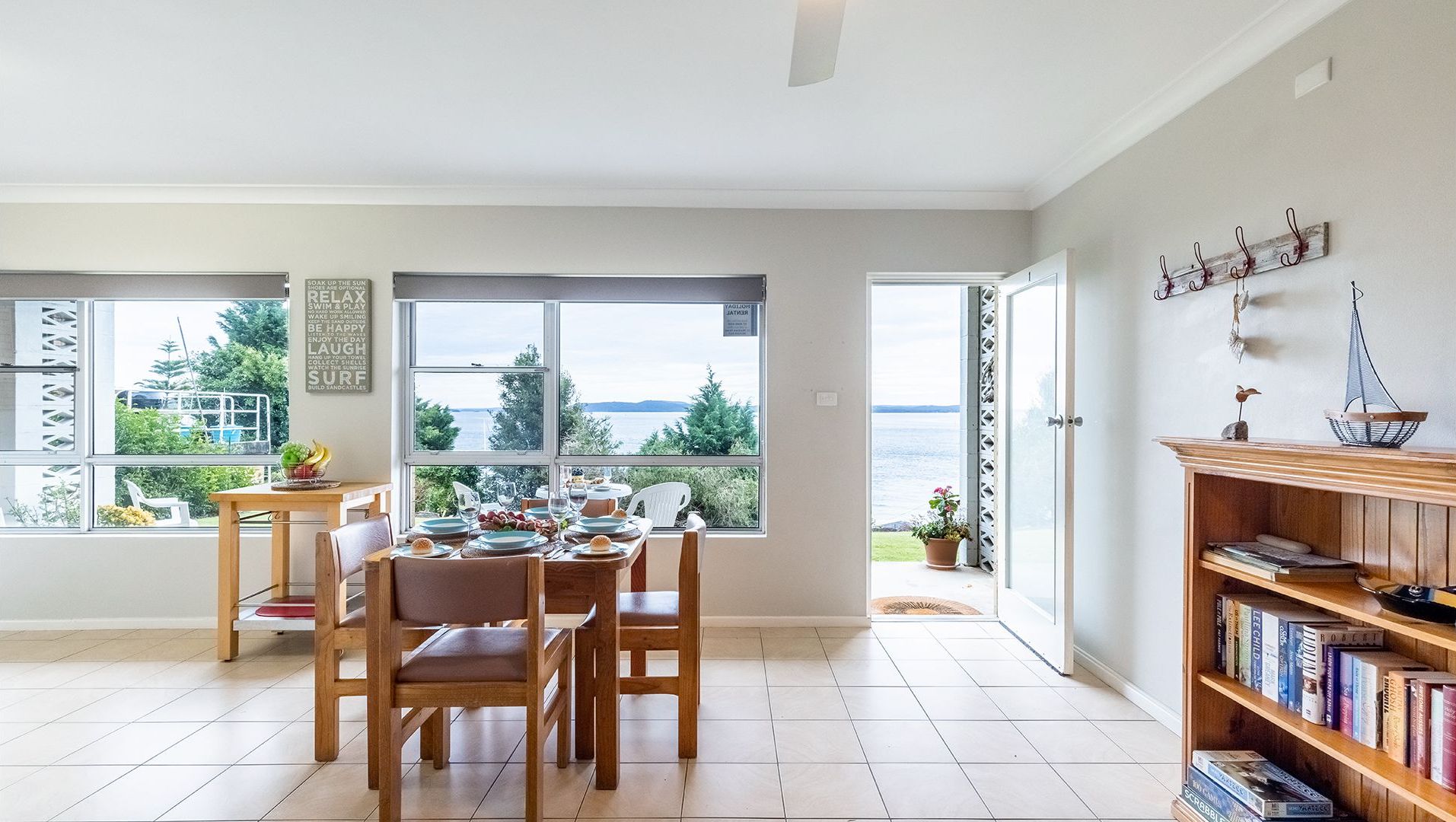 A dining area with a table set for four, large windows offering a view of the sea, and a bookshelf filled with books.
