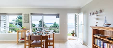 A dining area with a table set for four, large windows offering a view of the sea, and a bookshelf filled with books.