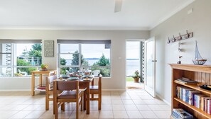 Un coin repas avec une table dressée pour quatre personnes, de grandes fenêtres donnant sur la mer et une bibliothèque remplie de livres.