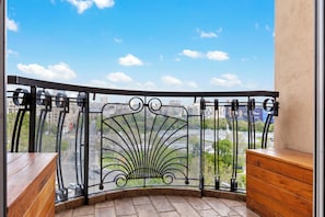 A balcony with a curved metal railing, a wooden bench, and a view of a cityscape with buildings and greenery.