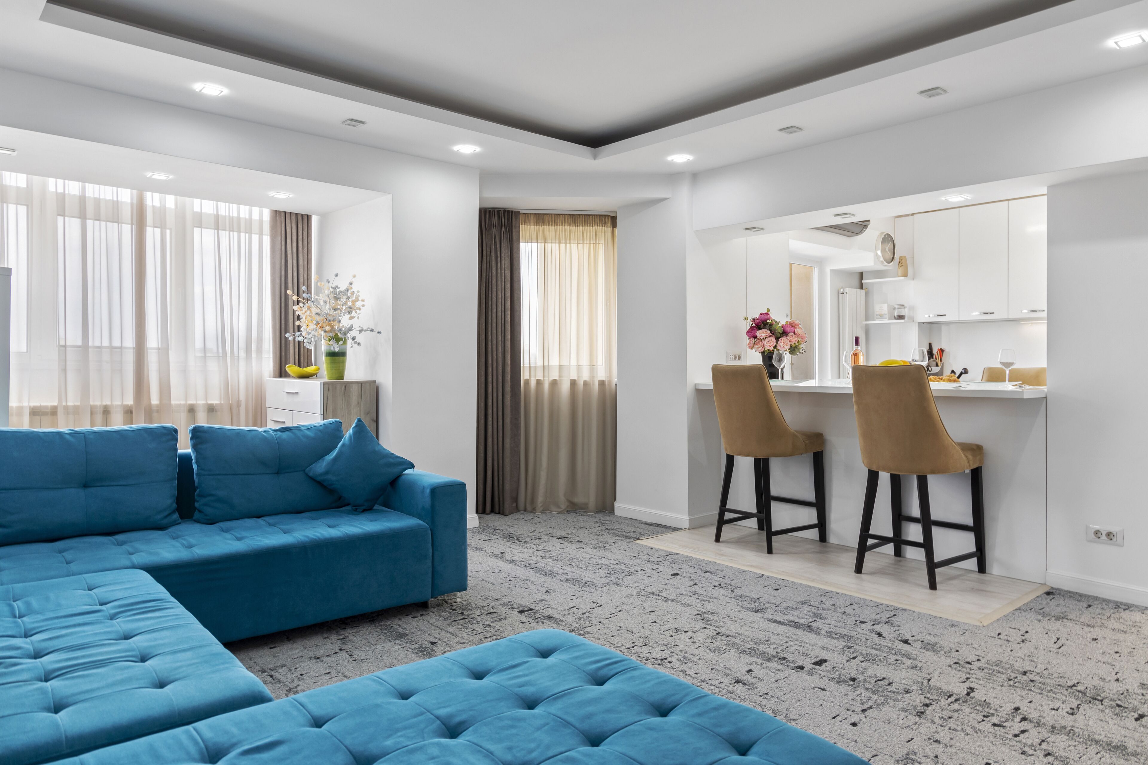 A modern living room with a sectional sofa, a dining area with bar stools, and a kitchen with white cabinets.