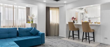 A modern living room with a sectional sofa, a dining area with bar stools, and a kitchen with white cabinets.