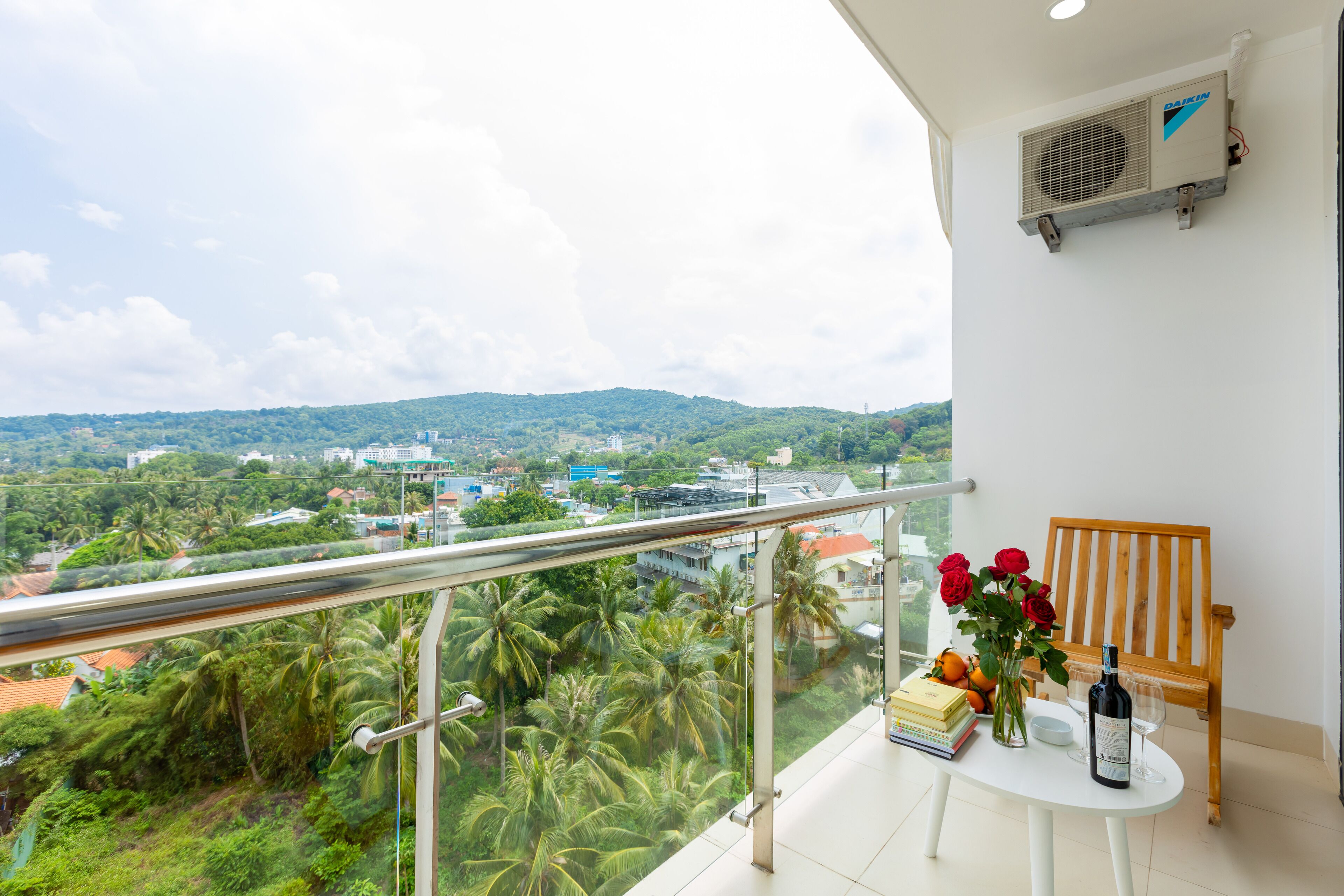 A balcony with a view of a cityscape and greenery, a table with books, a bottle, and a vase of flowers.
