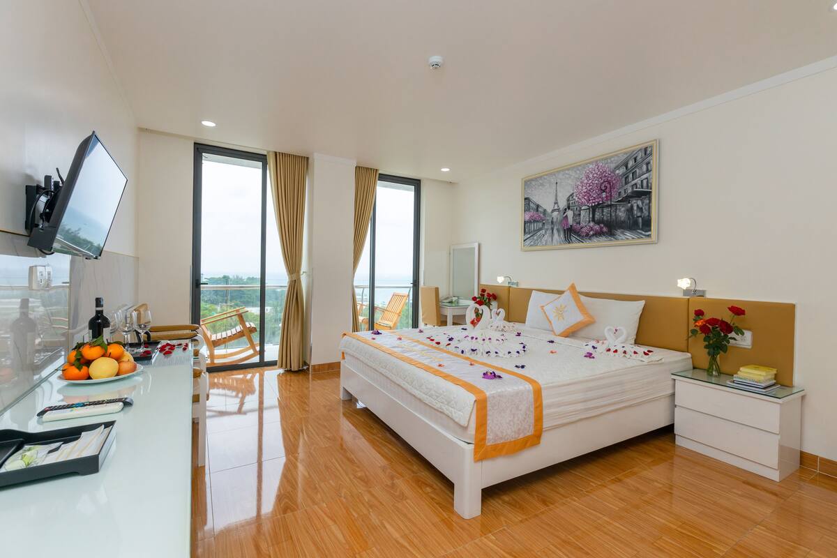 A modern hotel room with a large bed, a flat-screen TV, a dining area with a table and chairs, and a balcony with a view.