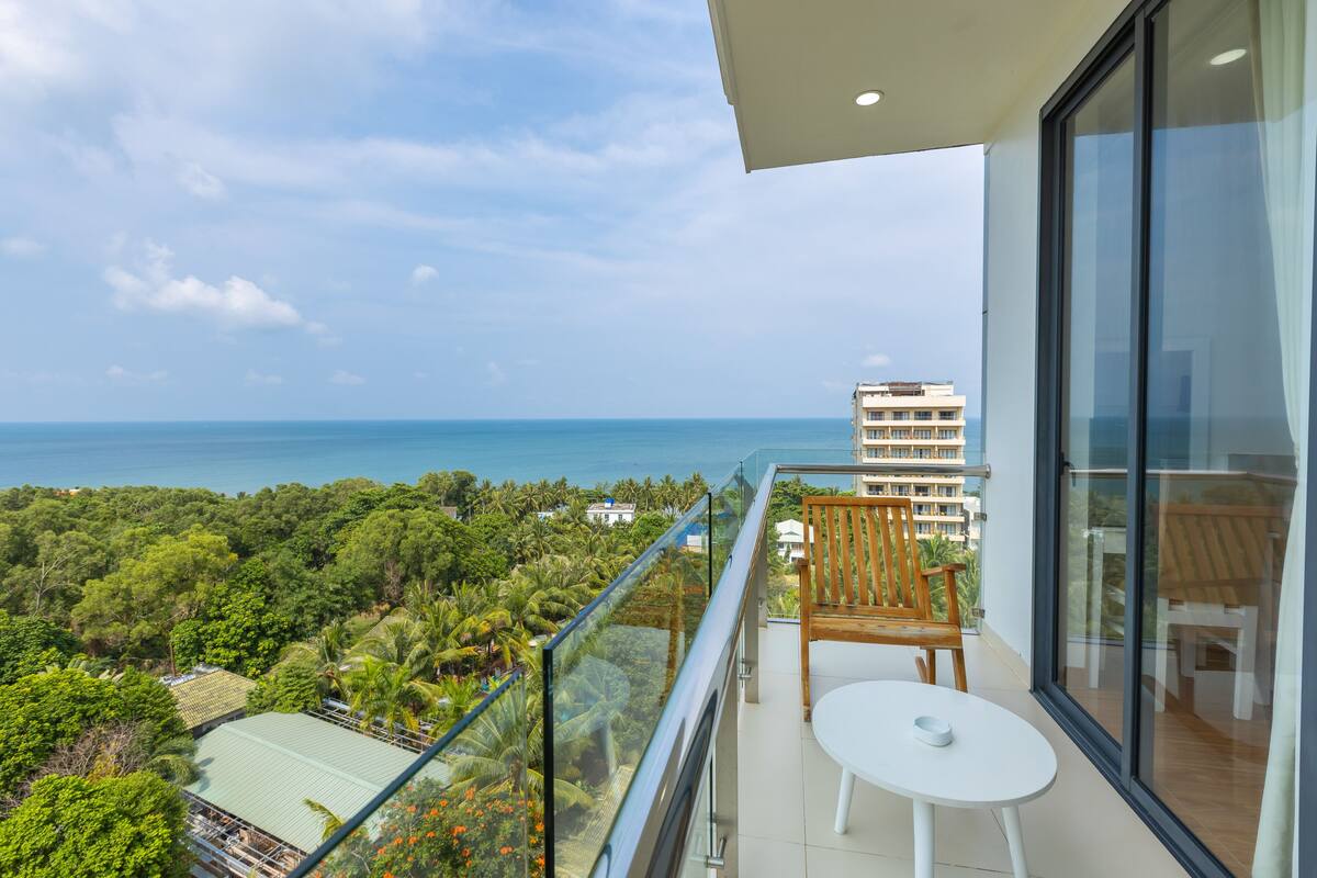 A balcony with a glass railing, a wooden chair, a small round table, and a view of the ocean and surrounding greenery.