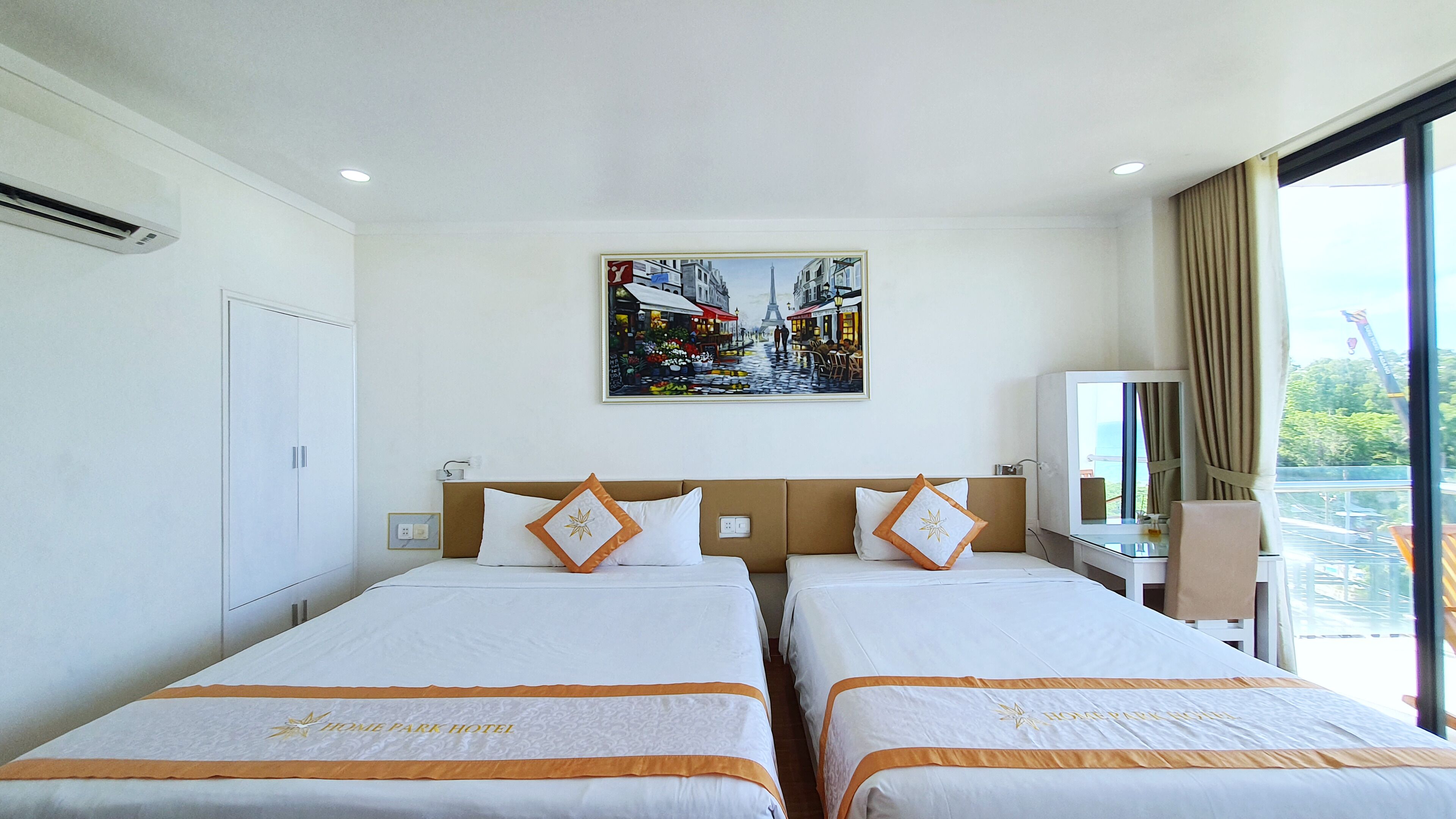A hotel room with two beds, a painting of a city street, a desk, and a view of the outdoors.