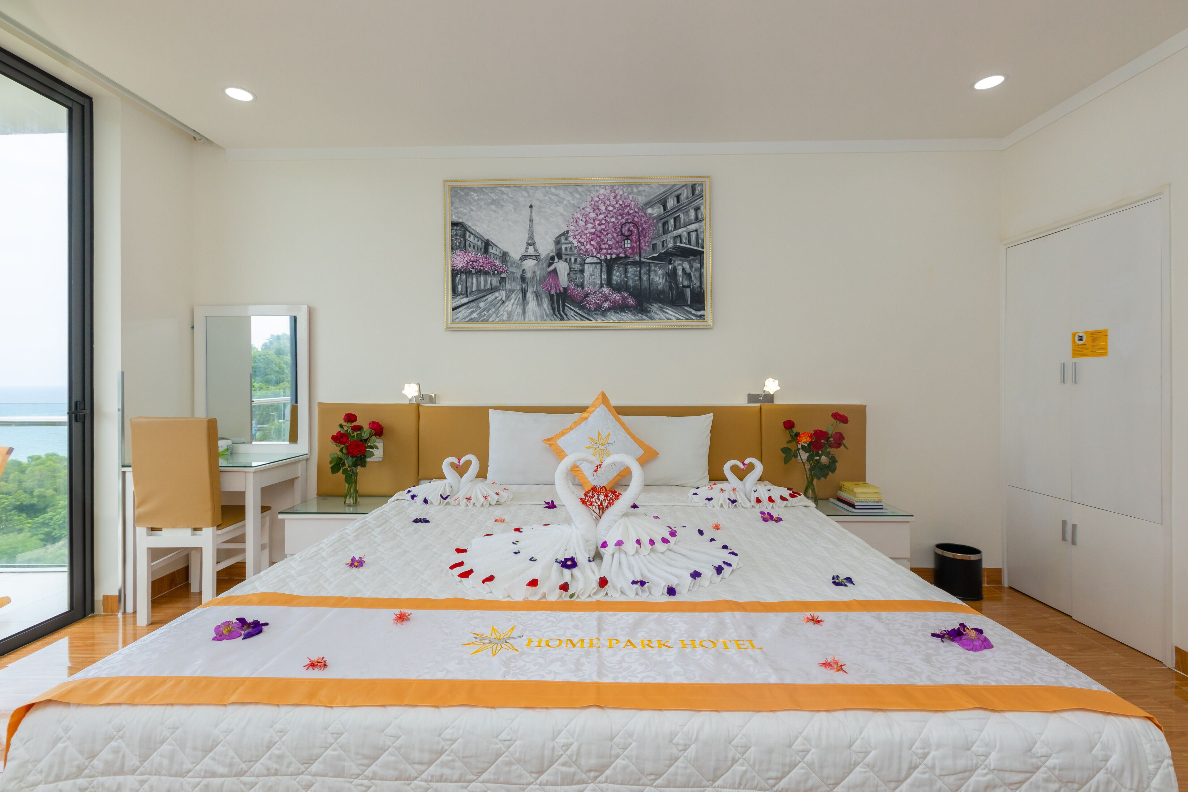 Habitación de hotel con una cama decorada con un cisne y un arreglo floral, un escritorio con una silla, un espejo y una pintura de un paisaje urbano.