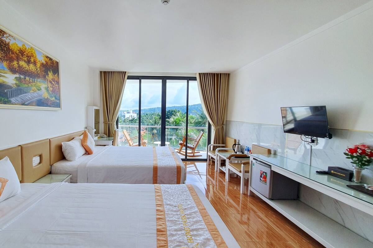 A hotel room with two beds, a desk with a TV, a balcony with a view, and a painting on the wall.