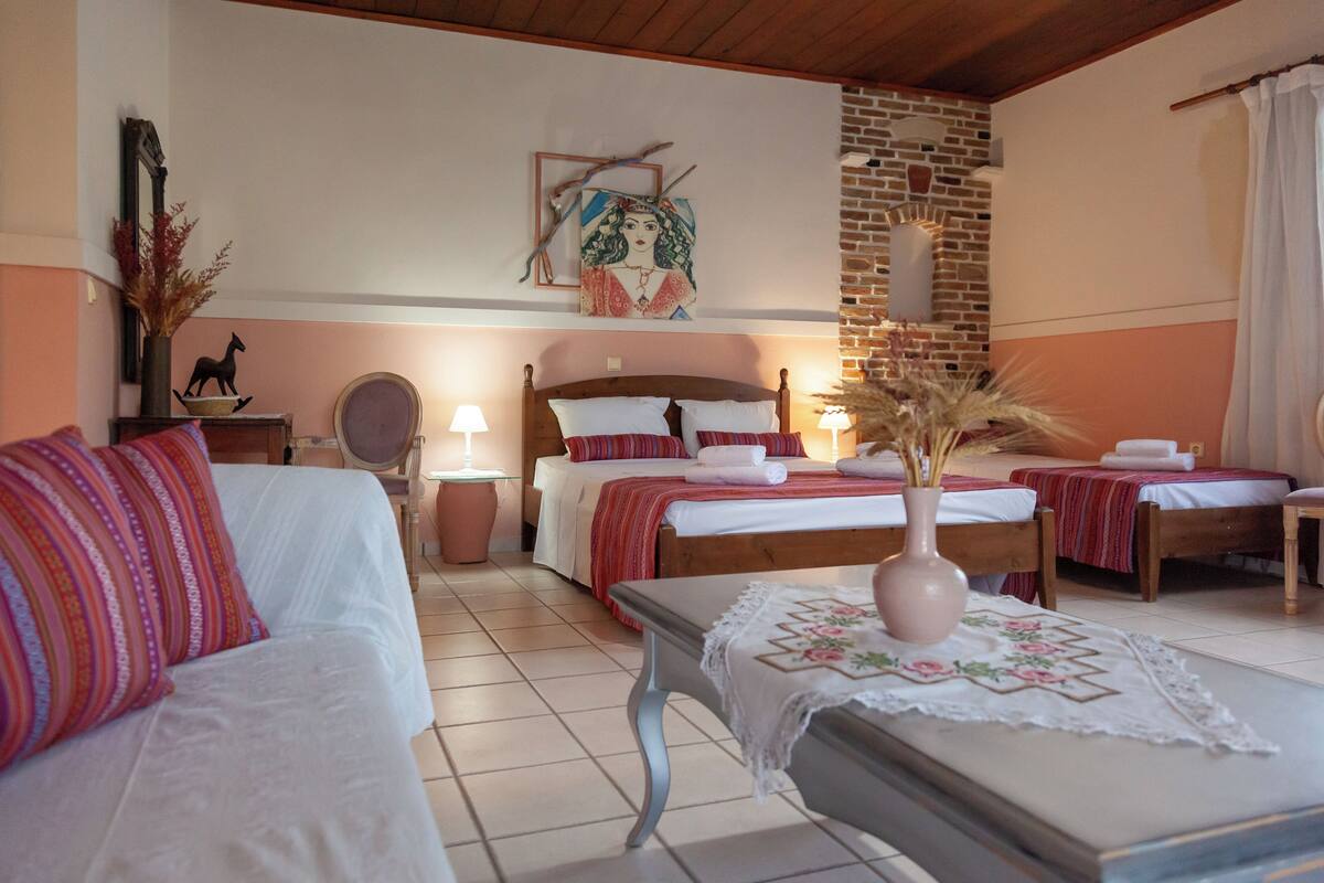 Quarto de hotel com uma cama grande, duas camas menores, um banco de madeira, um vaso com plantas secas, um espelho e uma obra de arte na parede.
