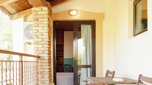 A covered outdoor patio with a wooden table and chairs, a stone wall, and a view into a room with a bed and curtains. - Amnisiades Apartments (Heraklion)