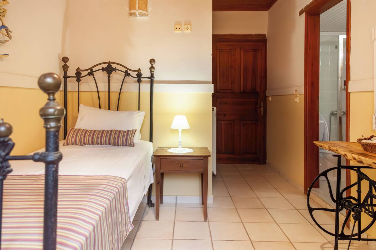 Quarto com cama, mesa de cabeceira com abajur, guarda-roupa de madeira e uma pequena mesa com cesta.