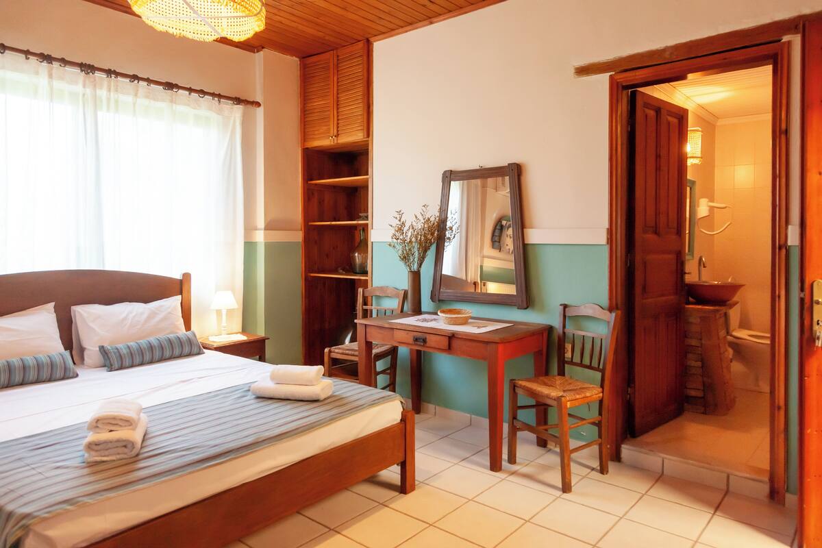 Quarto de hotel com cama, escrivaninha, cadeira, espelho e banheiro visível por uma porta aberta.