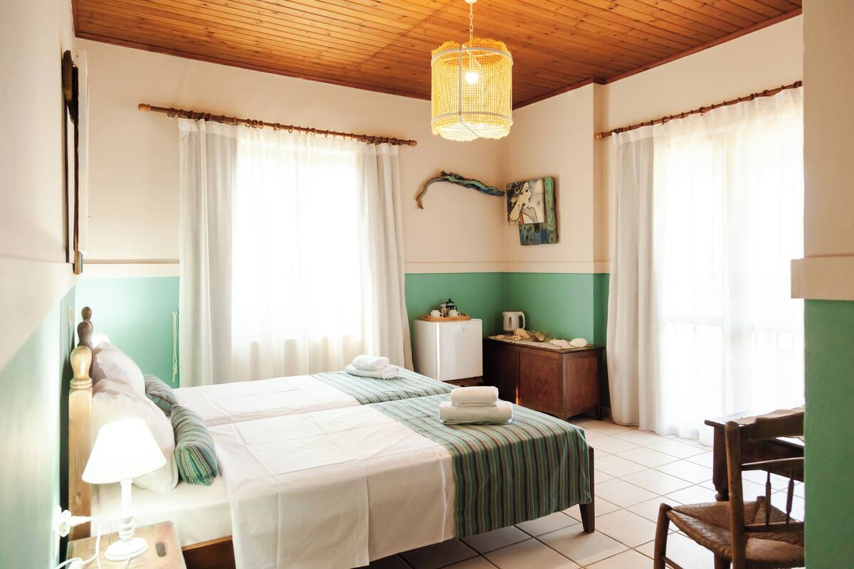 Quarto com duas camas, teto de madeira, luminária pendente, mesa pequena com abajur e uma cadeira.