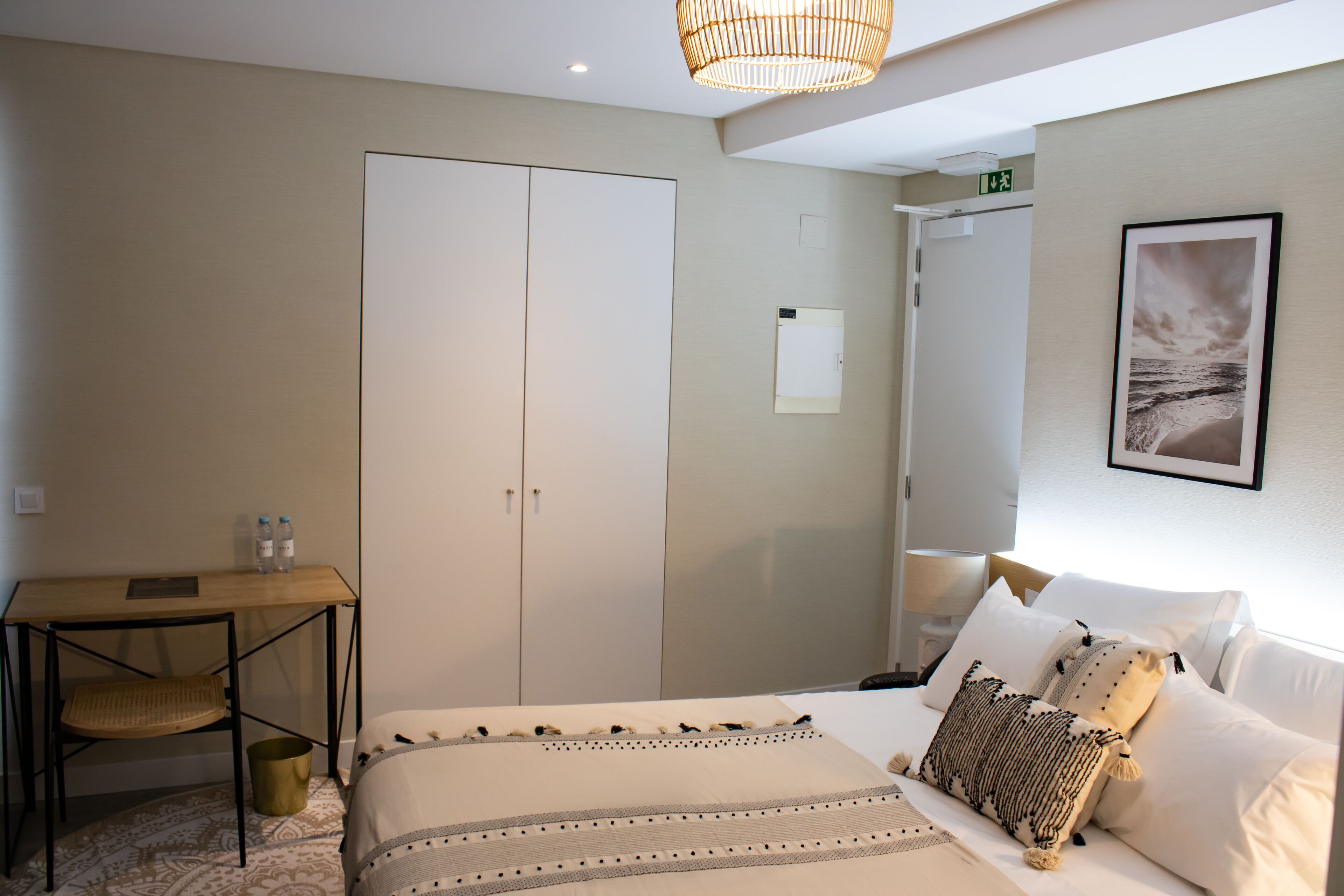 A hotel room with a bed, bedside table, lamp, wardrobe, and a framed picture on the wall.