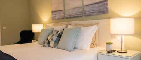 A neatly made bed with a white comforter and multiple pillows, a bedside table with a lamp, a small potted plant, and a remote control.