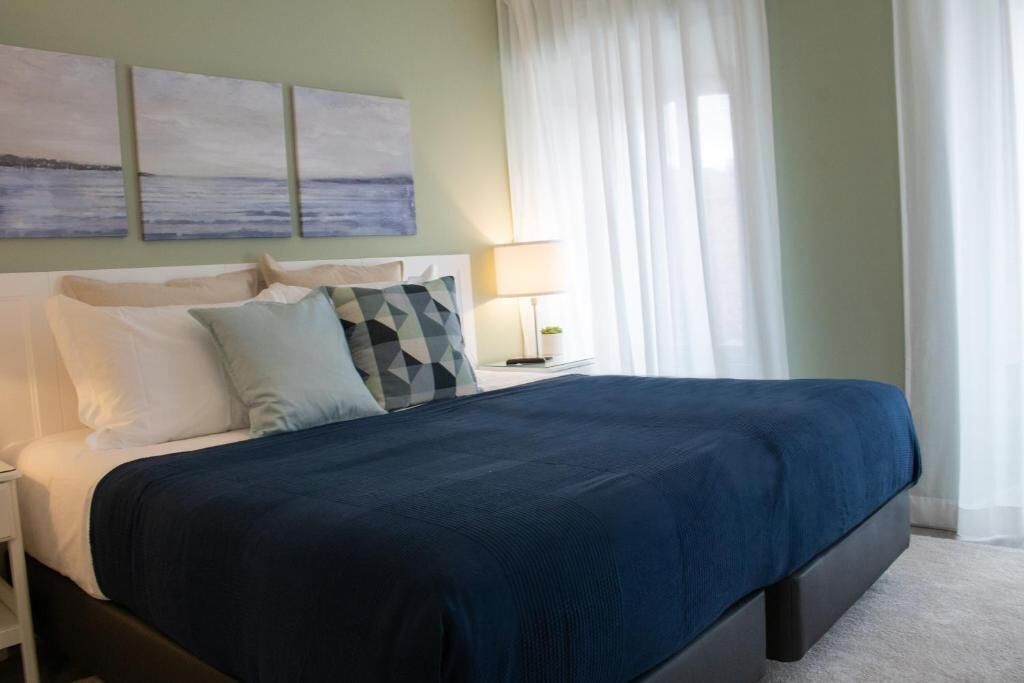 Una cama bien hecha con una colcha azul oscuro y varias almohadas, una lámpara de noche y tres cuadros colgados en la pared.