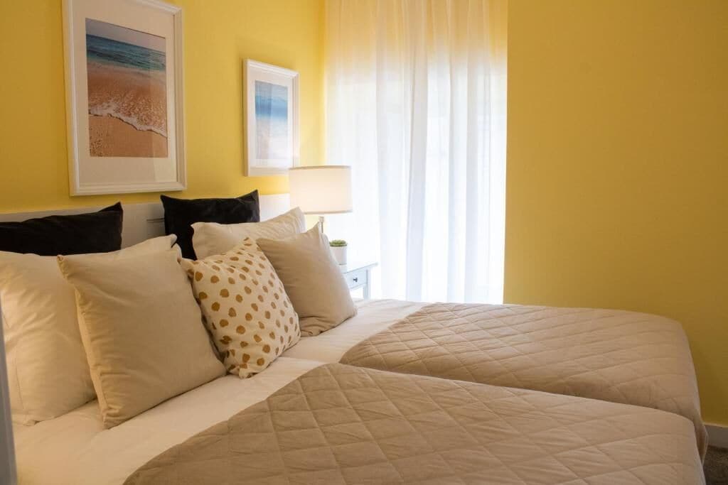 Cama doble con ropa de cama color crema, mesita de noche con lámpara y dos cuadros en la pared.