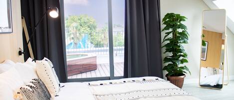 A modern bedroom with a large bed, a potted plant, a mirror, and a view of the outdoors.
