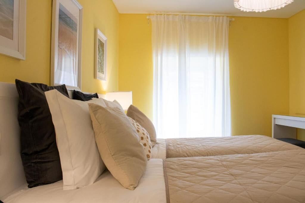 A room with yellow walls, a bed with beige bedding, and a window with white curtains.