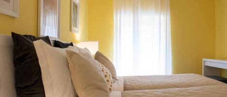 A room with yellow walls, a bed with beige bedding, and a window with white curtains.