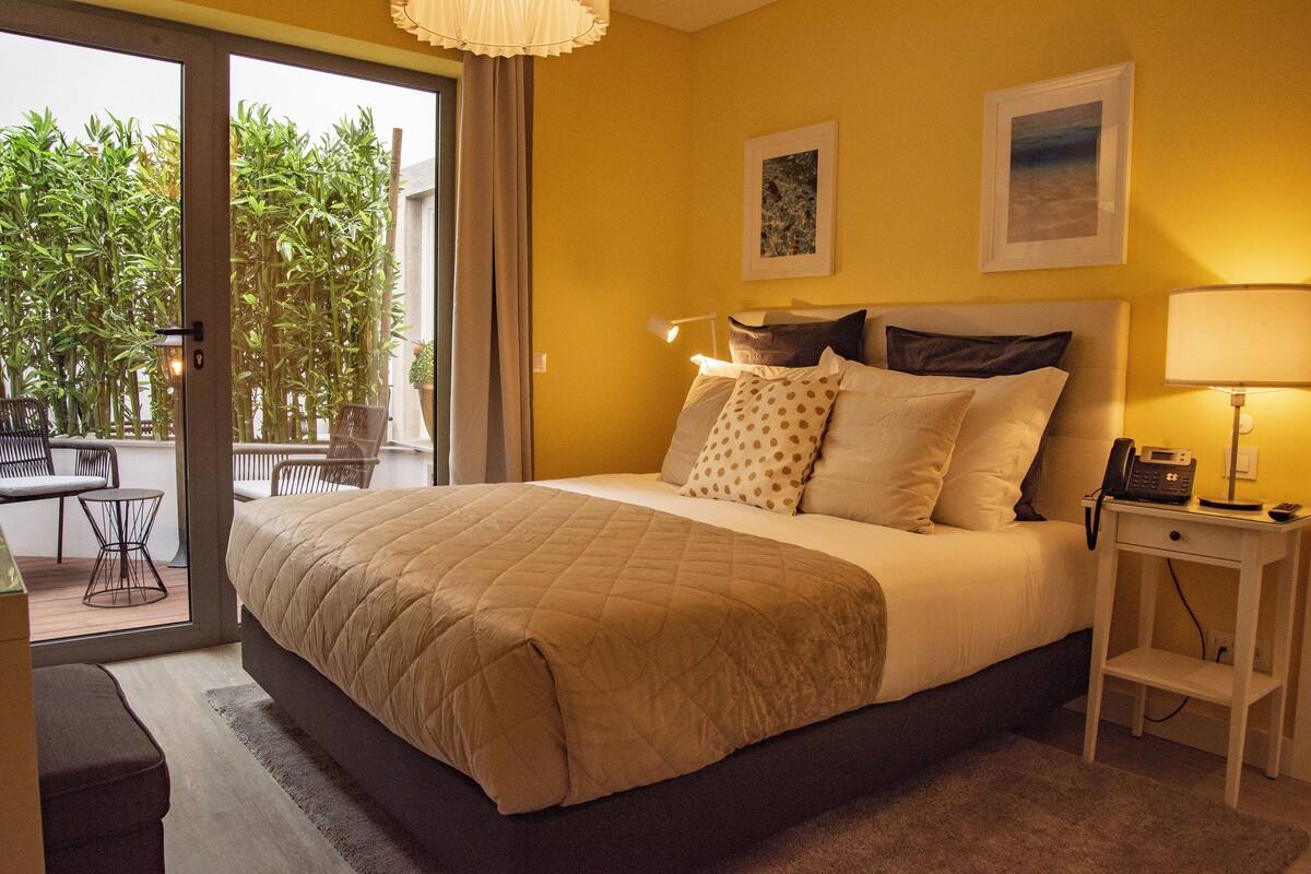 A bedroom with a large bed, a bedside table with a lamp, and a view of an outdoor area with chairs and a table.
