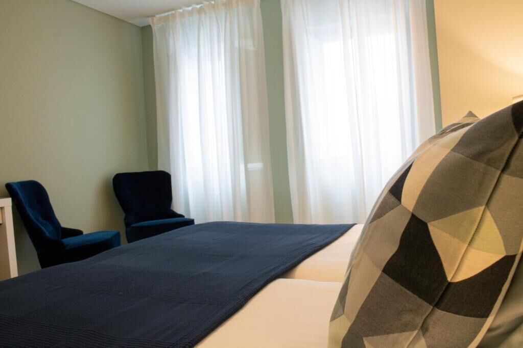 Habitación de hotel con una cama, dos sillas y un ventanal con cortinas translúcidas.