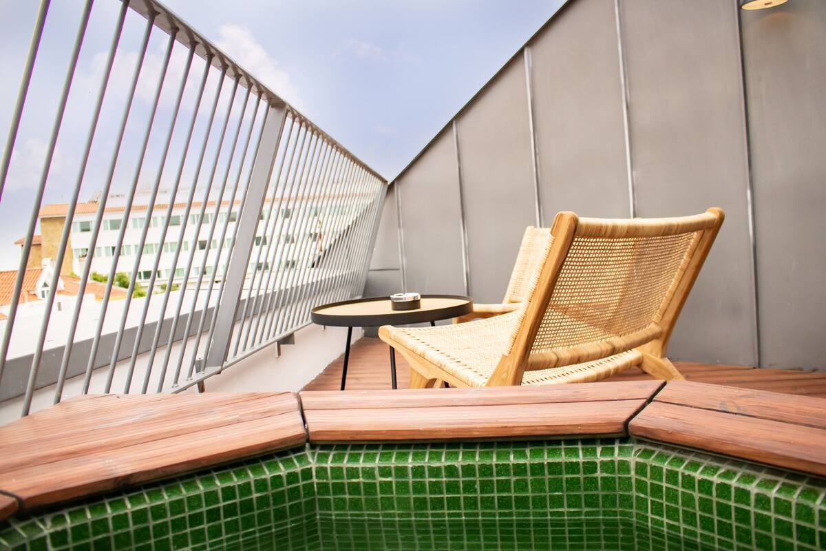 A balcony with a wicker chair and a small table, a green tiled area, and a view of buildings in the background.
