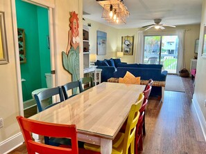 Uma sala de jantar com uma mesa de madeira e cadeiras coloridas, um sofá azul ao fundo e uma parede verde.