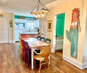 Uma sala de jantar com mesa de madeira e cadeiras coloridas, uma cozinha ao fundo e um grande painel na parede.